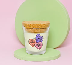 Container candle with cork lid. Candle has an orange heart with be mine, a purple heart with love and a pink heart with xoxo. It is on a green foam platform with a large round green circle behind it. The background is light pink.